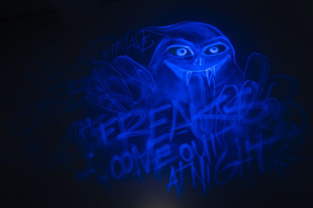 Auf einer schwarzen Wand erscheint in UV-Licht ein Monster mit grossen Augen und fletschenden Zähnen, unter dem steht Freak.