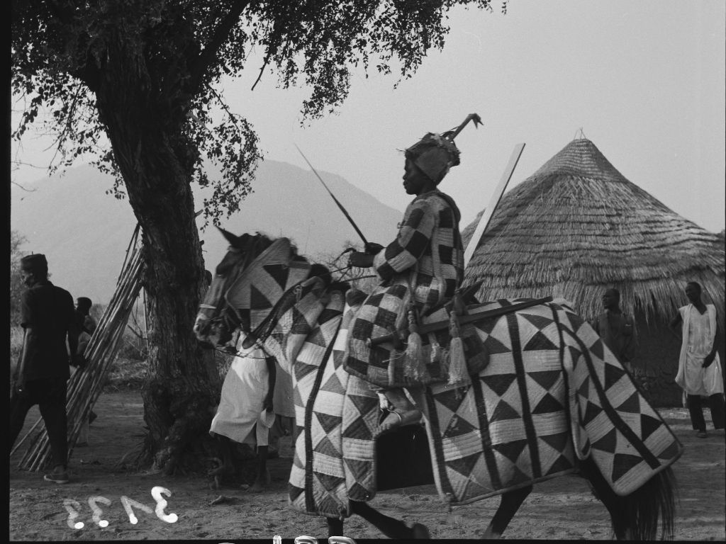 Schwarzweiss Fotografie von einem Reiter in einem dick wattierten Überzug, der sowohl Reiter als auch das Pferd bedeckt. Im Hintergrund ein grosser Baum und eine strohbedeckte Hütte.