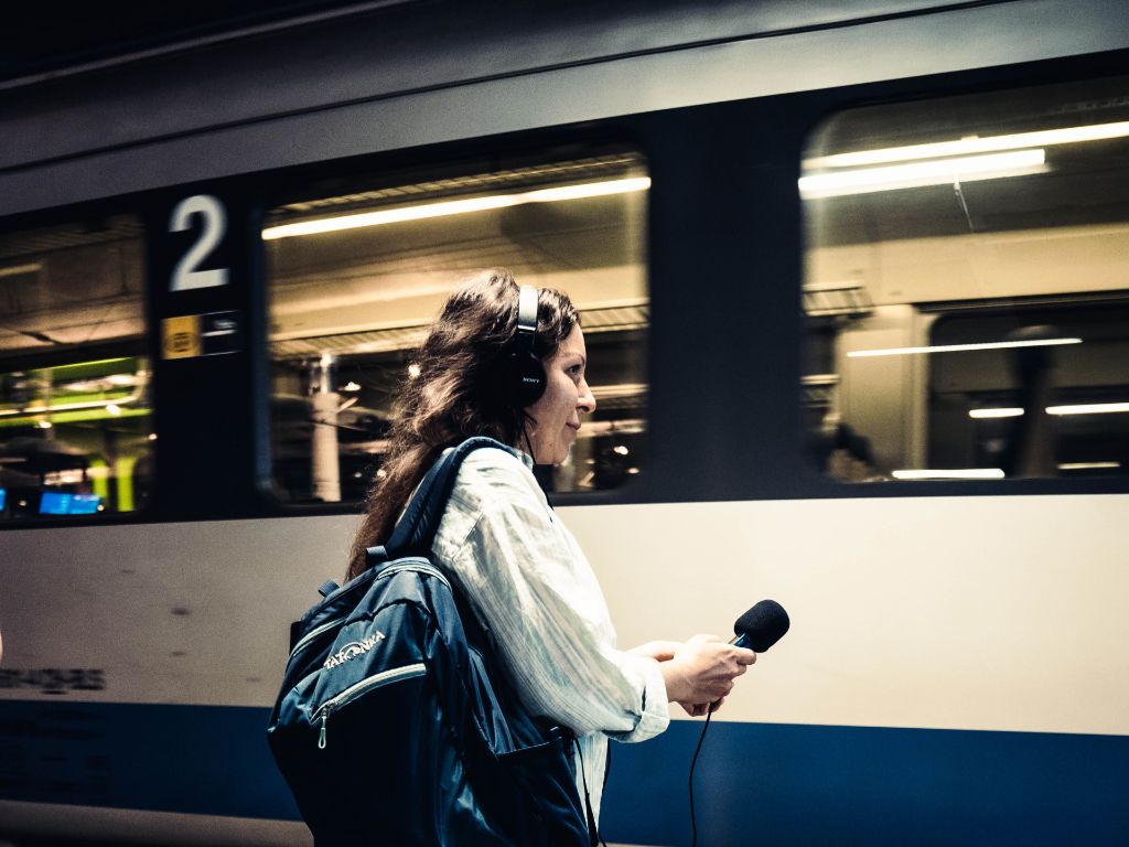 Frau von der Seite am Bahnsteig. Sie hat lange dunklen Haaren, trägt ein helles Hemd, einen grossen Rucksack, Kopfhörer, und hält Mikrofon. Im Hintergrund ein Zugwaggon.