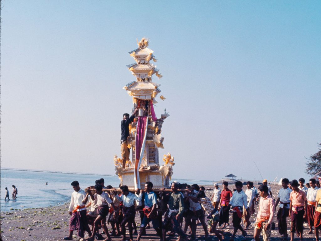 Fotografie von mehreren Menschen am Strand, die einen weiss-goldfarbenen, mit einem hohen Turm verzierten Sarg Richtung Wasser tragen.