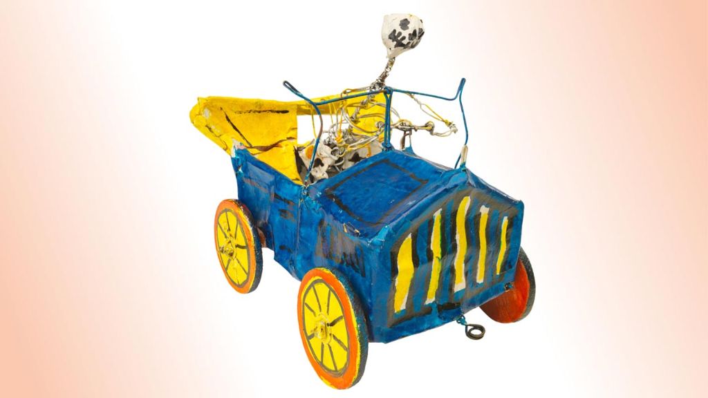 Mexikanische Totenfigur aus Pappmache, in Form eines Skeletts, welches ein blaues Oldtimer-Auto mit offenem gelben Verdeck, roten Rädern mit gelben Felgen steuert.