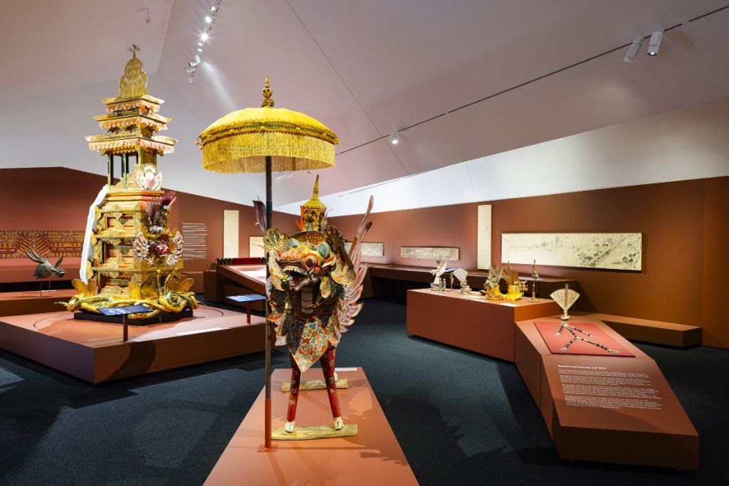 Blick in einen Ausstellungsraum mit roten Podesten, dunkelblauem Teppich und Objekten auf den Podesten und an den Wänden. Prominent zu sehen ein Drache mit einem Schirm und ein grosser Kremationsturm.