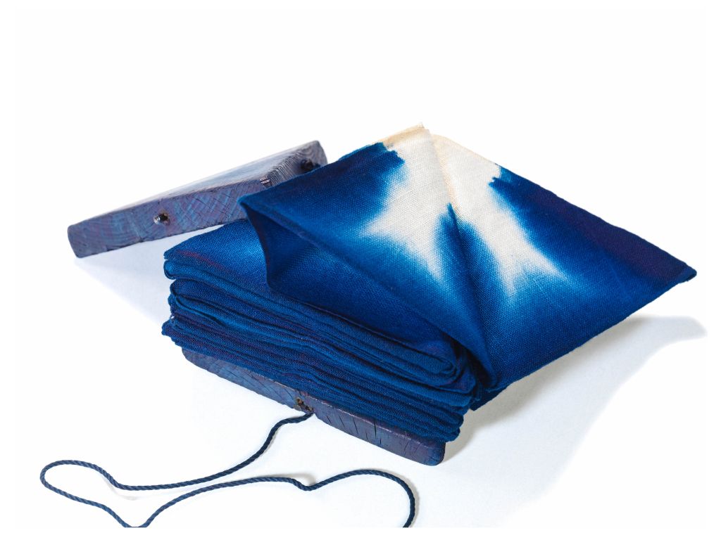Baumwollstoff (Oshime-Tuch), zwischen zwei dreieckigen, blau verfärbten Holzbrettchen gepresst. Der Stoff wird gefaltet, zwischen die Brettchen gepresst und mit Schnur fixiert.
