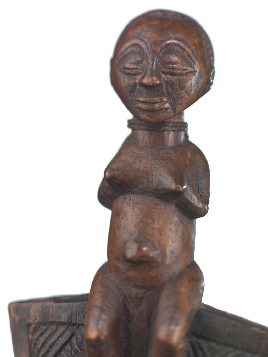 Eine Holzfigure sitzt auf einem Zeremonialstab. Es handelt sich um eine Frau. Sie sitzt aufrecht und hat die Hände über der Brust gefaltet. Sie hütet Geheimnisse.