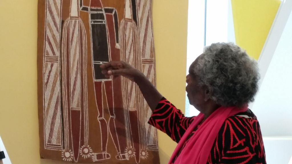 Ruth Nalmakarra steht vor einer Rindenmalerei und zeigt mit der rechten Hand auf Details