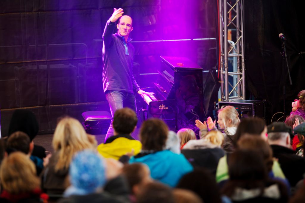 Auf dem Foto ist Andrew Bond im Zentrum auf der Bühne zu sehen. Er steht vor dem Klavier. Mit der einen Hand spielt er, mit der anderen lädt er das Publikum offensichtlich zum Mitsingen ein.