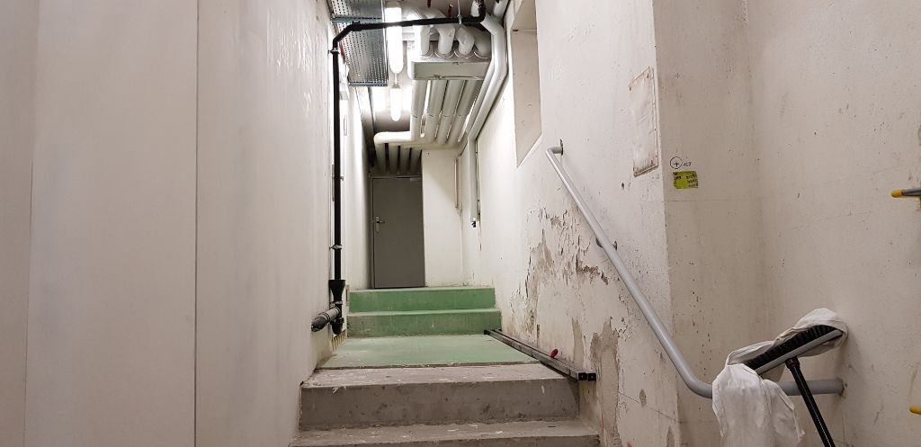 Ein enger Gang mit Treppenstufen führt zum Nachbarsgebäude. Die weissen Wände blättern stückweise ab, an der Decke hängen Rohre.