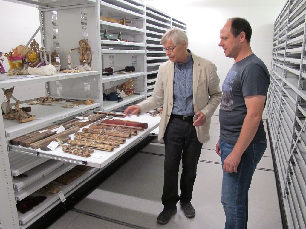 Werner Gamper steht neben Kurator Richard Kunz zwischen grauen Gestellen und zeigt auf eine offene Schublade mit Objekten aus seiner Sammlung