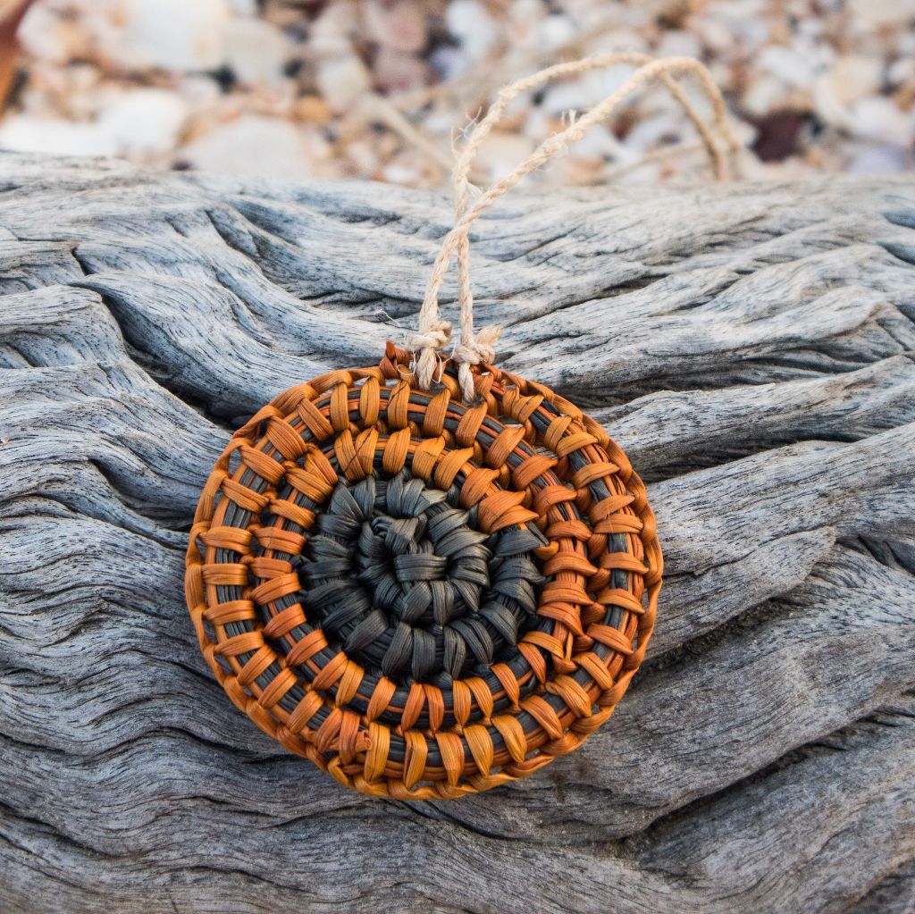 Ein geflochtener, runder Halskettenanhänger, einer Schnecke ähnlich, in orange und schwarz, liegt auf einem Baumstamm