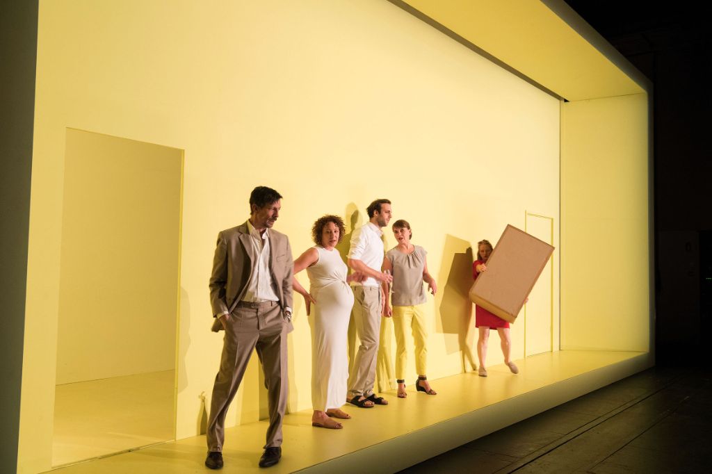 Auf dem Foto ist eine Szene aus einem Theaterstück des Theater Basel zu sehen. Mehrere Personen stehen in Reih und Glied auf der gelben Bühne. Ein Mann hebt sich ab respektive steht etwas abgedreht da.