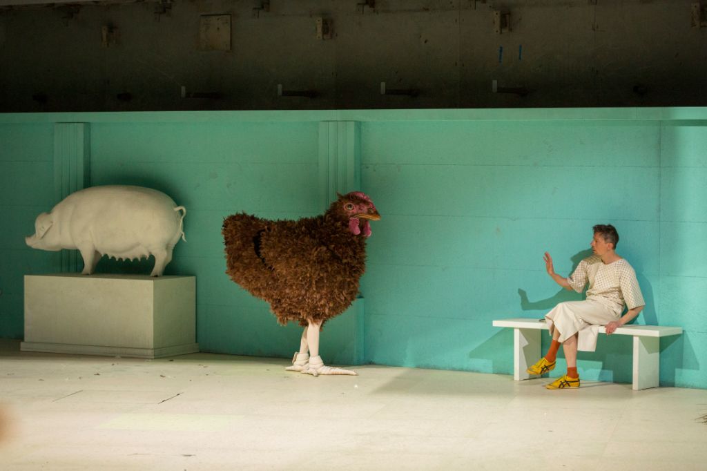 Das Foto zeigt eine Szene aus einem Stück des Theater Basel. Ein Mann sitzt auf einer Bank, neben ihm ein riesiges Huhn, das ihn anschaut. Neben dem Huhn steht ein Schwein auf einem Podest, da in die andere Richtung blickt.