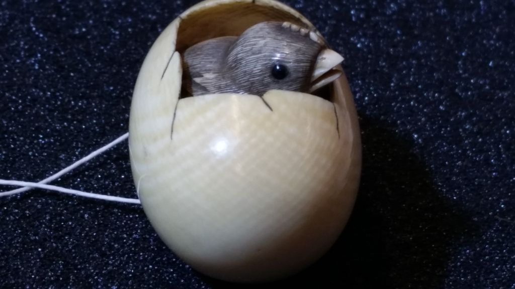 Zu sehen ist ein Ei aus Elfenbein. Es ist aufgeschlagen und darin sitzt ein Vögelchen. Das Elfenbein weist schraffierte bräunliche Linien auf.