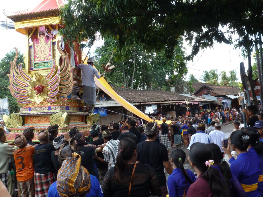 Eine Menschenmenge trägt einen Sarg durch ein Dorf, der wie eine Tempel aussieht