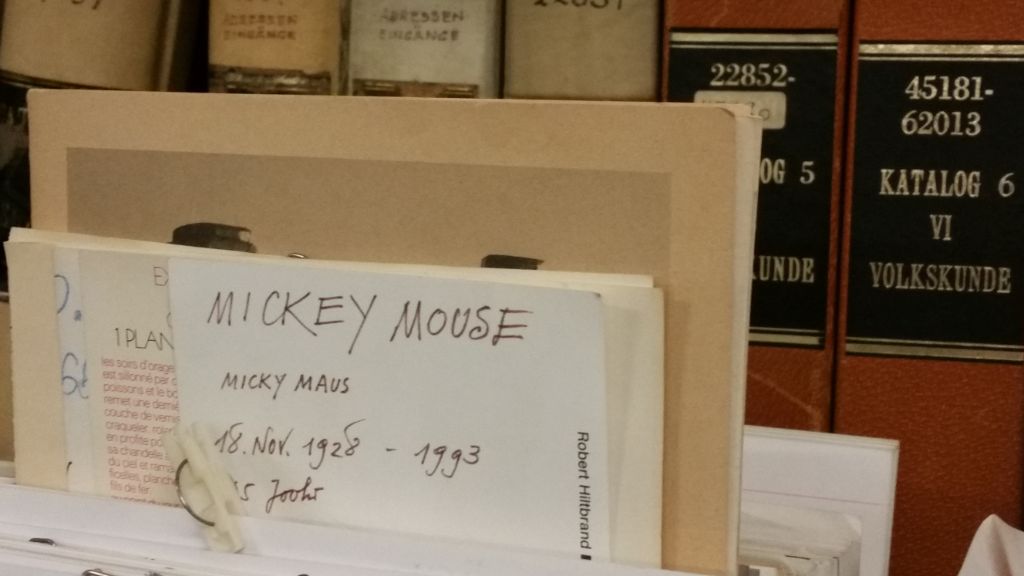 Das Foto zeigt eine Schachtel auf einem Gestell in einem unserer Depots. Daraus schaut eine Karte, auf dieser steht: Mickey Mouse und Micky Maus, 18. November 1928 ...