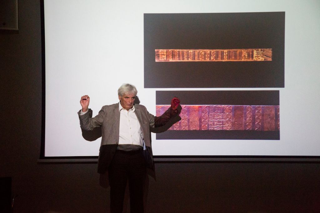 Vor einer Leinwand, auf der zwei Stoffmuster gezeigt werden, steht Professor Reimar Schefold mit erhobenen Armen und doziert