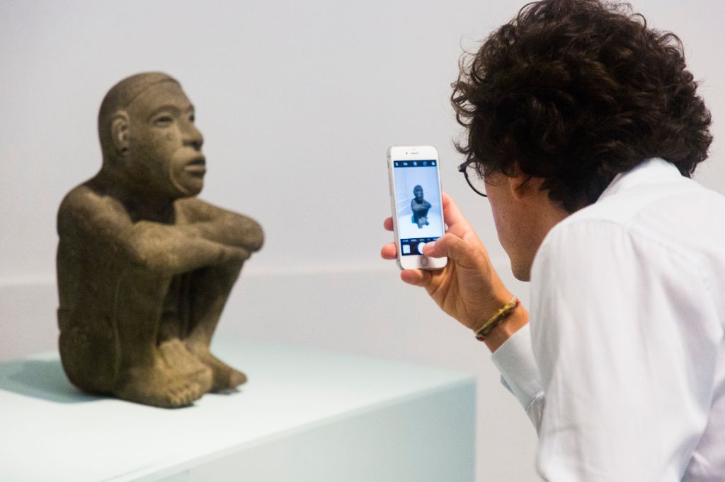 Ein Mann steht vor einer auf einem Podest sitzenden kleinen Figur und fotografiert sie mit seinem Handy.