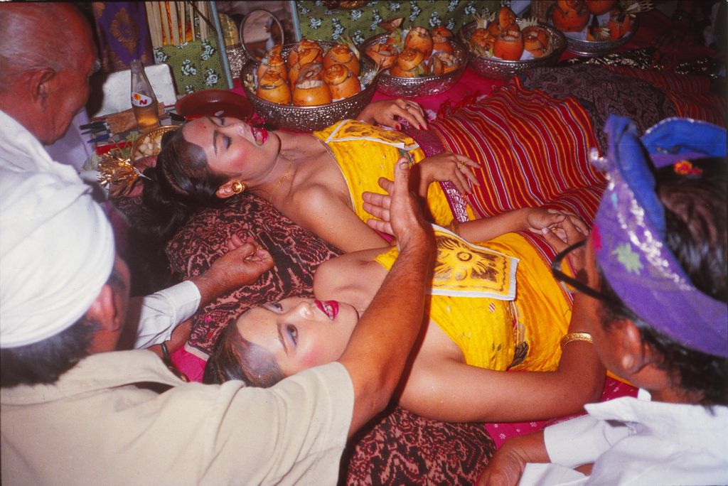 Zwei Frauen liegen auf Teppichen am Boden mit geöffneten Mündern. Sie sind in gelbe Gewänder gekleidet und schön geschminkt.  Ein Mann macht sich daran, ihre Zähne zu feilen.