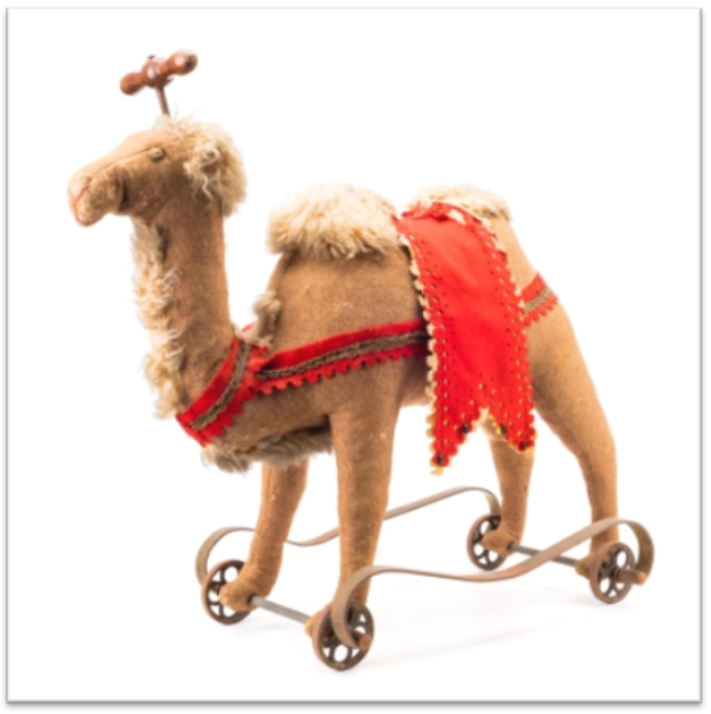 Das Kamel ist braun mit helleren Haaren. Es steht auf Rädern und hat auf dem Kopf einen Griff. Die Sitzfläche ist mit rotem Stoff ausgelegt, ebenso die Zügel.