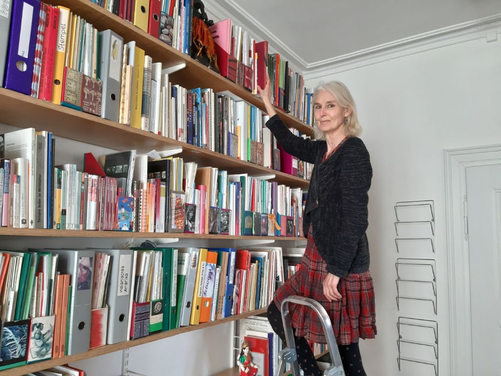 Regina Mathez, gekleidet in dunklen Pulli und kurzen Rock steht auf einer Leiter vor einer Wand mit Büchergestellen. Diese sind voll mit Ordnern.
