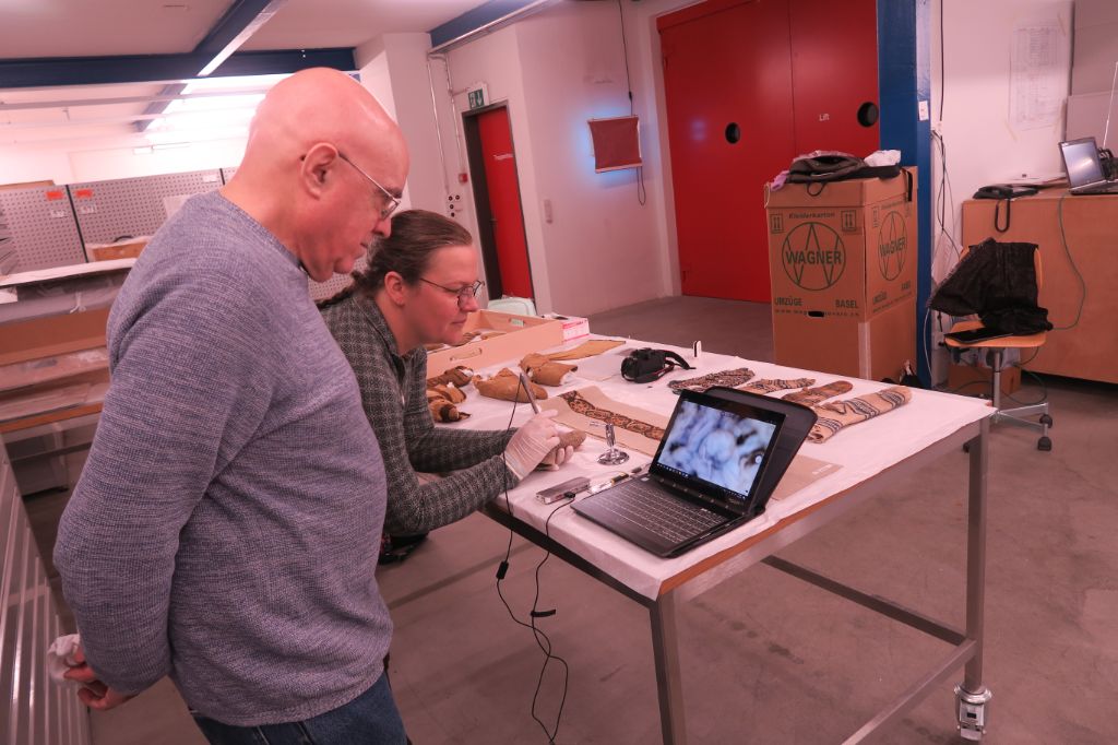 Auf dem Bild stehen Anne Marie und Cary vor einem Laptop, das auf einem Tisch im Depot steht. Sie schauen sich darauf vergrösserte Bilder an von einem Gegenstand, den Anne Marie in der Hand hält.