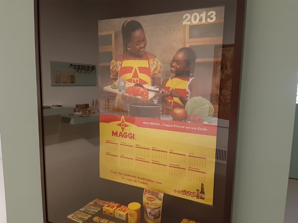 Das Foto zeigt einen Maggi-Kalender aus dem Jahr 2013. Unten ist ein Kalender in gelb und rot, darüber ist ein Foto zu sehen, das eine Mutter und eine Tochter in einer Küche zeigt. Sie tragen beide gelb-rote Maggi-Kochschürzen und schauen auf verschiedene Esswaren.