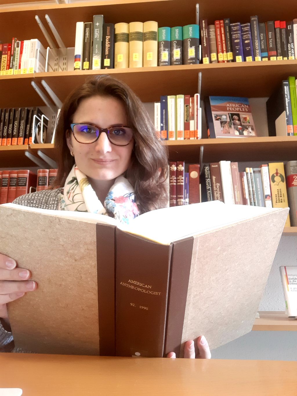Das Foto zeigt Viktoria Terendi in unserer Bibliothek, vor einer Wand voller Bücher. Sie sitzt an einem Tisch, hält ein grosses und dickes Buch in der Hand. Viktoria hat langes braunes Haar und trägt eine Brille.
