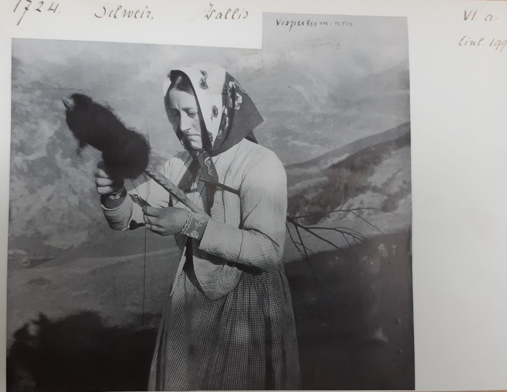 Es ist ein altes Schwarz-Weiss-Foto zu sehen. Im Zentrum steht eine Frau mit Rock, Strickjacke und Kopftuch. Sie hält eine Art Spindel in der Hand und unter dem Arm einen kleinen Ast eingeklemmt. Hinter ihr sieht man schneebedeckte Berge.