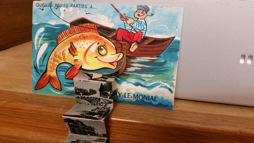 Das Foto zeigt eine gemalte Postkarte, auf der das Meer zu sehen ist. Darauf ein kleines Boot mit einem Fischer. An der Angel hat er einen riesigen Fisch. Aus dem Bauch des Fisches entrollen sich Schwarz-Weiss-Fotos der Ortes, von wo die Karte kommt.