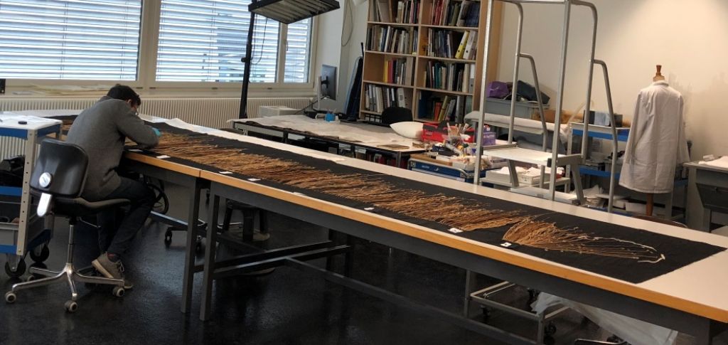 Das Foto zeigt Medrano sitzend an einem langen Tisch. Darauf ausgebreitet ist das fünf Meter lange Stück Stoff mit den Schnüren. Medrano beugt sich über den Stoff. Im Hintergrund ist das Restaurierungsatelier zu sehen.