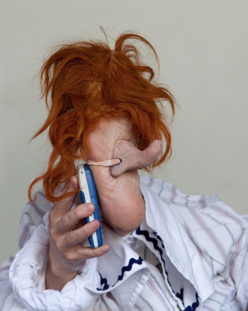 Auf dem Foto ist eine Fussfigur zu sehen, sie trägt wuschelige rote längere Haare, hat eine lange gebogene Nase und telefoniert mit einem Handy