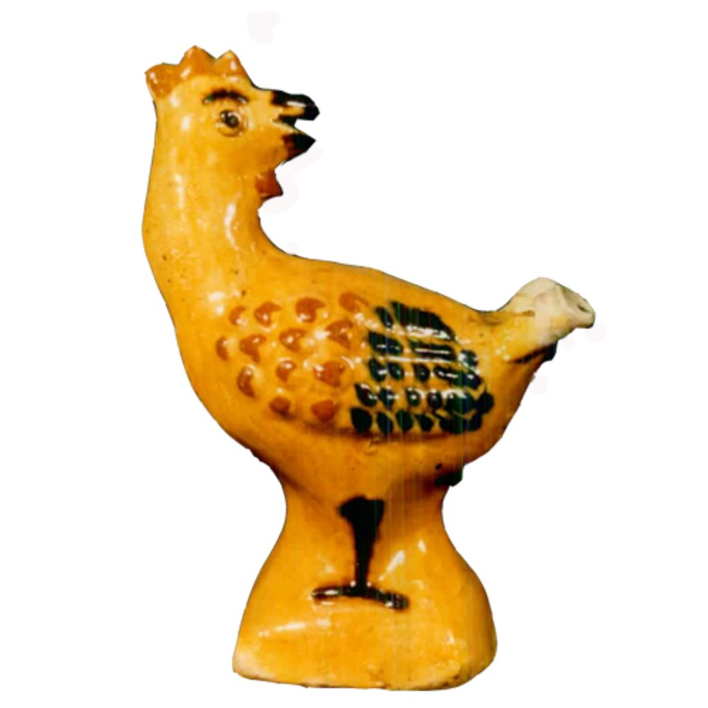 Das Foto zeigt einen Hahn aus Keramik. Er ist ockerfarben sowie rot und schwarz bemalt. Der Kopf zeigt nach hinten zum Schwanz.