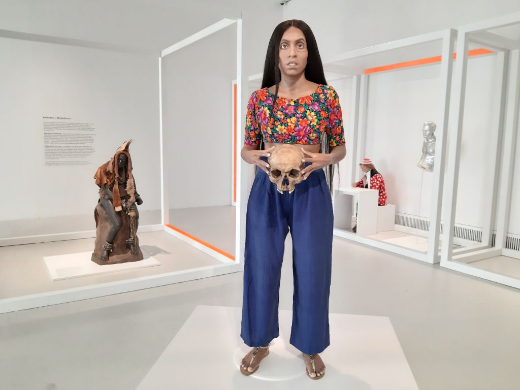Das Foto zeigt die Figur der Künstlerin auf einem weissen Podest in der Mitte eines weissen Ausstellungssaals. Sie trägt blauen Hosen und ein farbiges Top. In den Händen hält sie einen Schädel.
