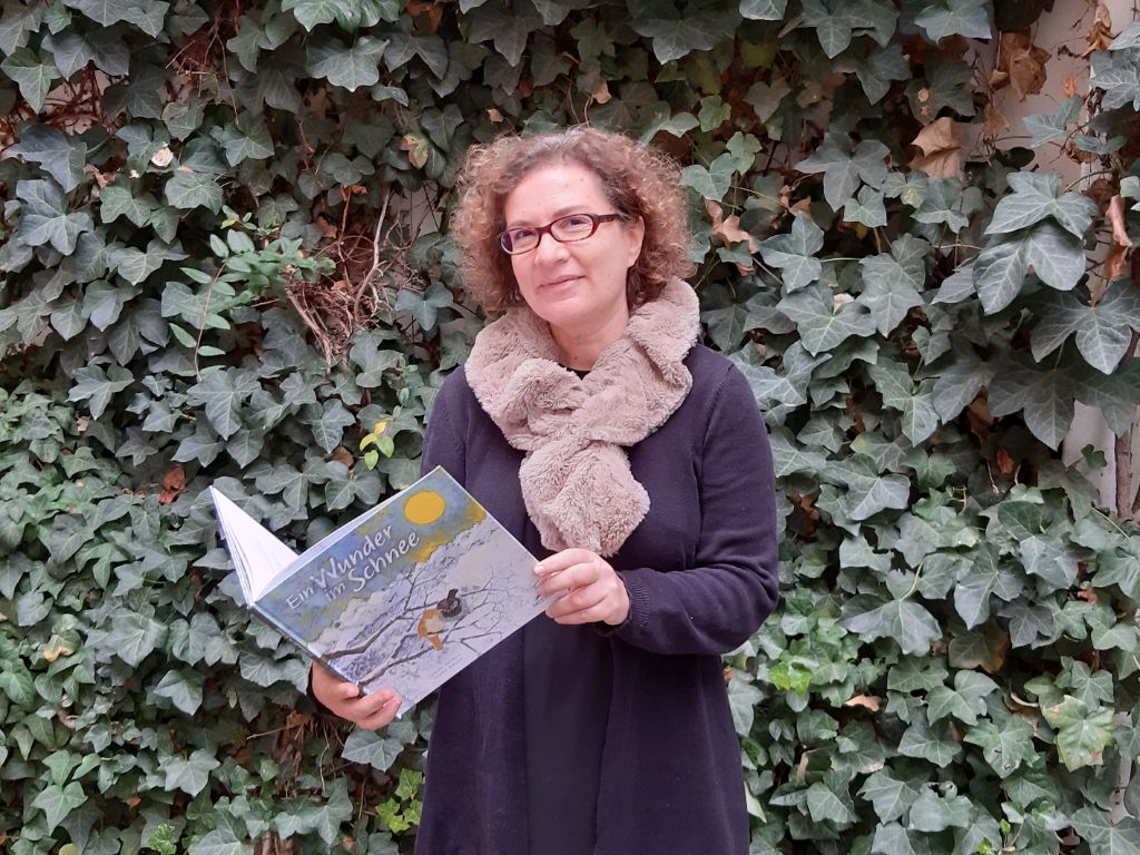 Sabine Rotach steht in blauer Strickjacke mit beigem dickem Schal vor einer Wand aus Efeu und hält lächelnd ihr Buch in der Hand