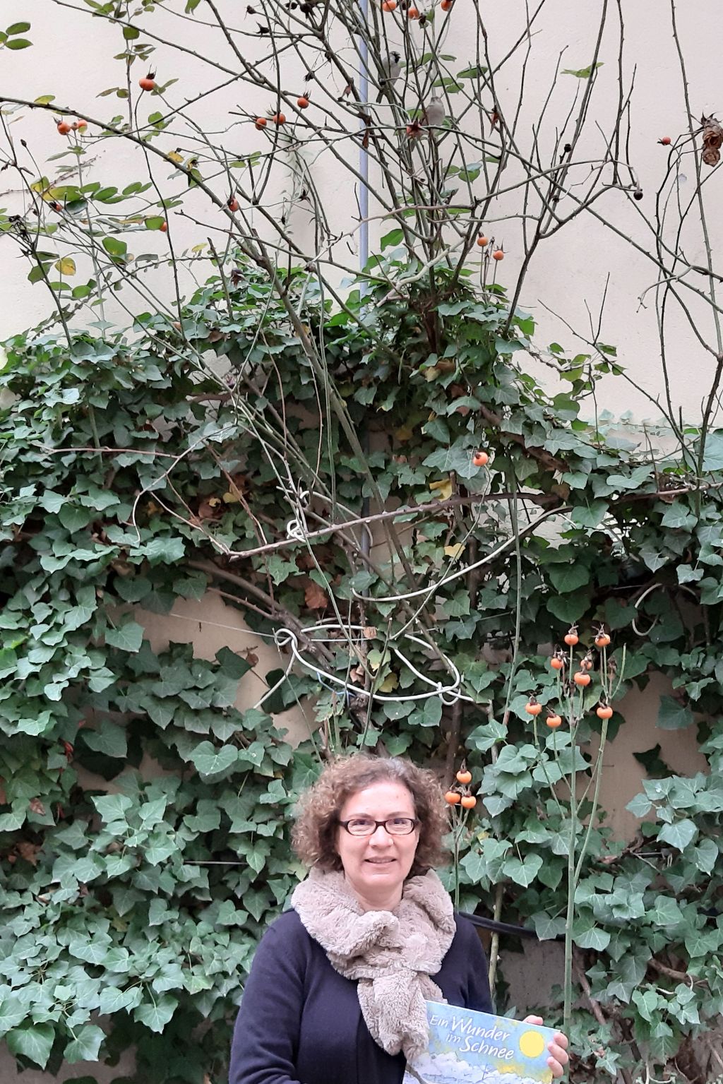 Sabine Rotach steht vor einer Wand, die mit Efeu bewachsen ist. Oben an ihr tummeln sich zwei Vögel in den Zweigen eines Baumes