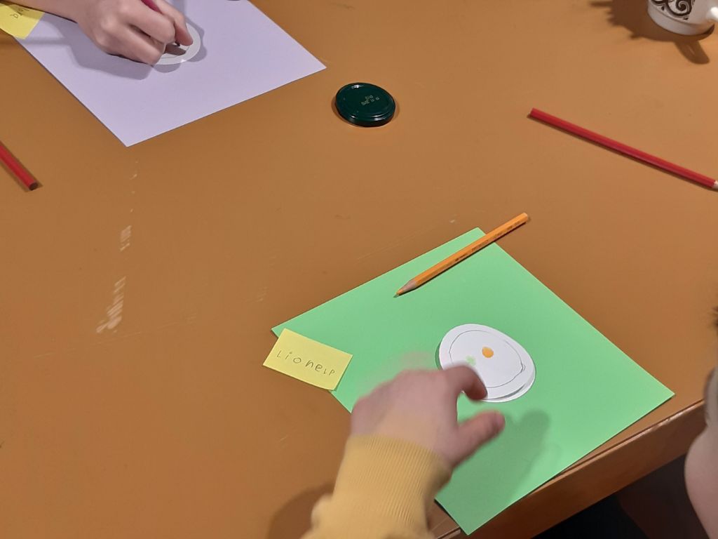 Das Bild zeigt einen Tisch, auf dem farbige Blätter liegen, auf denen die Kinder kleine weisse runde Blätter bemalen