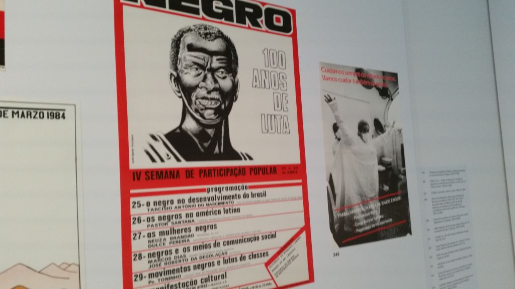Das Foto zeigt zwei Plakate, auf einem ist unter dem Titel Negro der Kopf eines Schwarzen und in der unteren Hälfte sind Daten aufgeführt. Das zweite Plakat daneben zeigt Pflegepersonal in Schutzkleidung