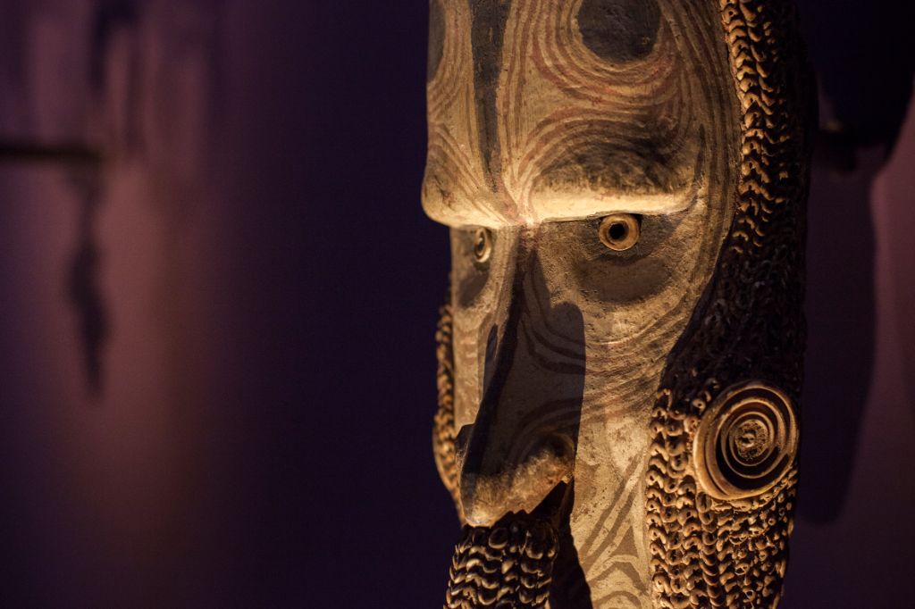 Das Foto zeigt eine längliche Maske aus Holz, die mit kleinen Muscheln verziert ist. Das Gesicht der Maske hat eine lange Nase und kleine, leuchtende Augen