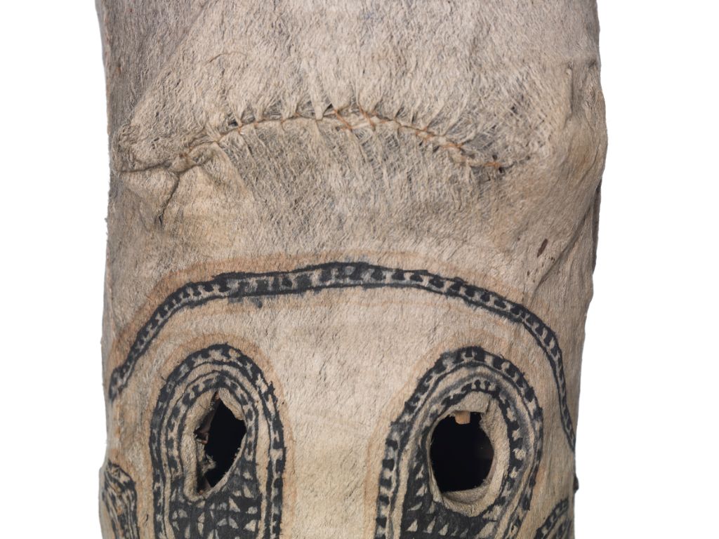 Auf dem Bild sieht man den oberen Teil einer Maske, das heisst nur die runden, ausgeschnittenen Augen und die Stirn. Die Maske ist eine längliche, weissliche Röhre. Augen und Stirn sind mit blauen Mustern verziert.