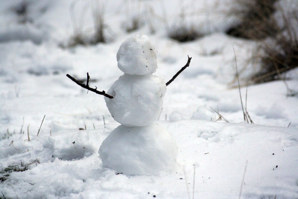ein kleiner Schneemann steht in einem Schneefeld, er besteht aus drei Kugeln und hat als Arme zwei Zweige