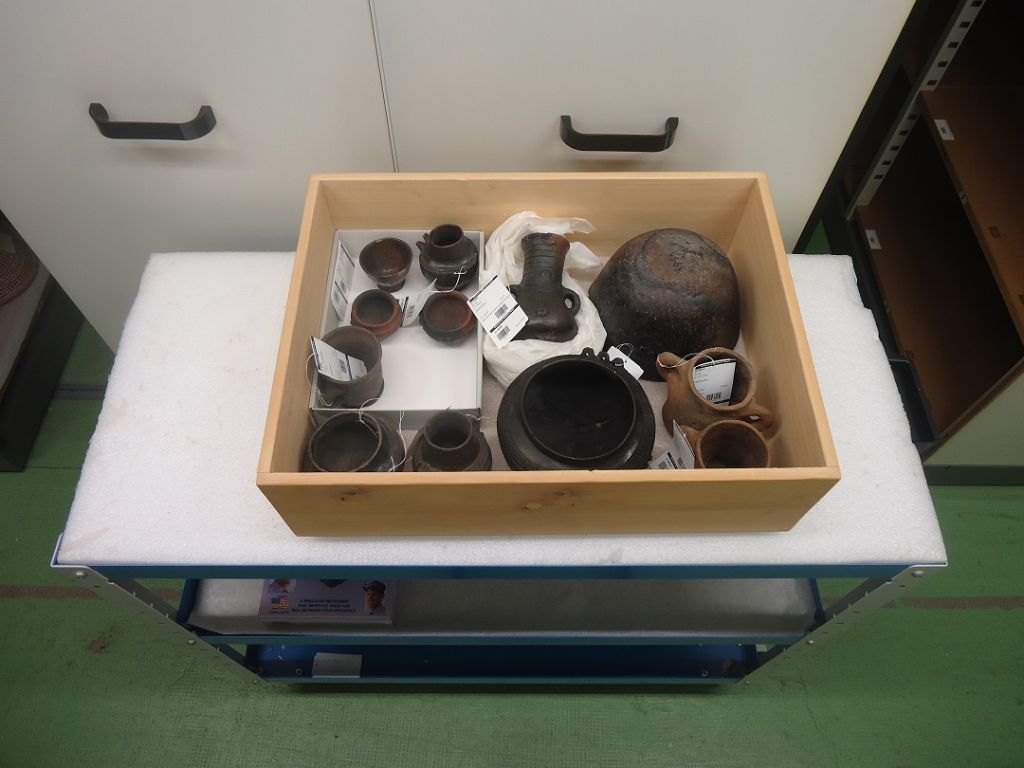 Holzkiste steht auf einem kleinen Tisch vor einem grauen Kasten. In der Kiste befinden sich dunkelbraune Gegenstände.