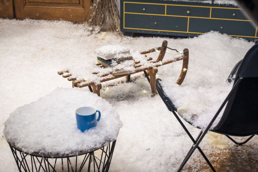 Links vorne steht ein kleine schwarzes Tischchen mit blauer Tasse, alles schneebedeckt. Daneben ein schwarzer Stuhl, ebenfalls voller Schnee. Im Hintergrund ein Schlitten mit Büchern, zugedeckt mit Schnee