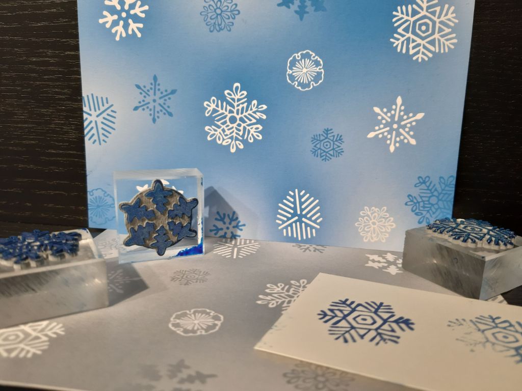 Blau-weisse Karte mit Schneeflockenmotiven stehend, davor liegend silbrig-weisse Karte mit Schneeflockenmotiven, darauf stehen und liegen drei Stempel mit Schneeflockenmotiven und ein weisses Kärtchen, auf dem die Stempel ausprobiert worden sind.