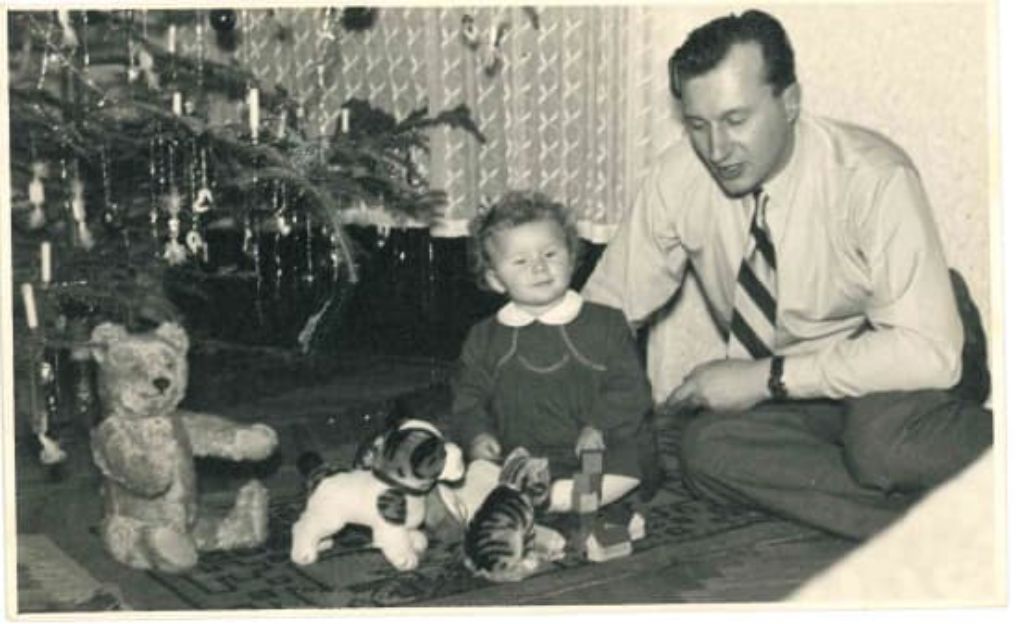 Unter einem geschmückten Weihnachtsbaum sitzt ein Teddybär. Daneben ein Plüschhündchen. Neben dem Spielzeug sitzt ein kleines Mädchen mit Kleid mit weissem Kragen. Neben ihr kauert ihr Vater im weissen Hemd mit gestreifter Krawatte.