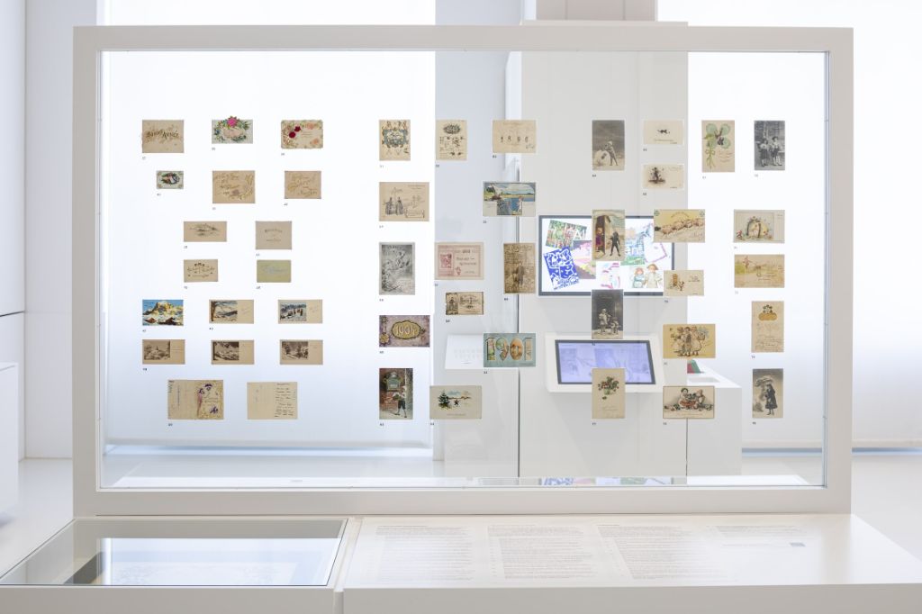 Auf einer transparenten Wand kleben winzig kleine Karten und Karten in Postkartenform. Durch die Wand hindurch sieht man zwei Bildhschirme.