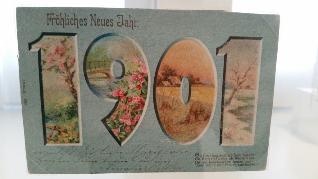 Bläuliche Postkarte mit der Jahreszahl 1901. In der 1 ist eine Frühlingslandschaft, in der 9 eine Sommerlandschaft, in der 0 eine Herbstlandschaft und in der 1 eine Winterlandschaft abgebildet. Darüber steht: Fröhliches Neues Jahr. Darunter ist von Hand etwas geschrieben, das kaum zu entziffern ist.