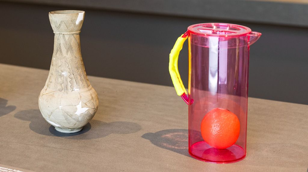 Links auf einem grauen Podest steht ein beiger Wasserkrug in Form einer Vase, unten bauchig, gegen oben schmaler, mit wenigen Verzierungen und weissen reparierten Stellen. Rechts von ihm steht ein pinkfarbener Plastikkrug mit Deckel, gelbem Griff und einer Plastikorange drin.