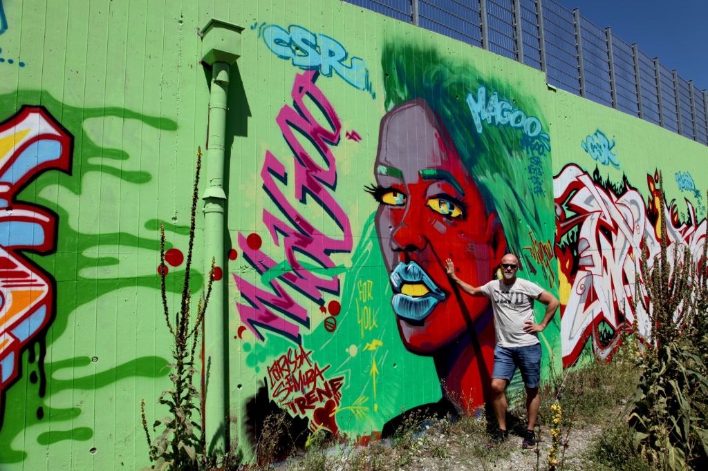 Magoo vor einem seiner neusten Grafftis am Tangentenweg in Basel. Zu sehen ein weibliches Gesicht mit grellen Farben, vorwiegend Rot.