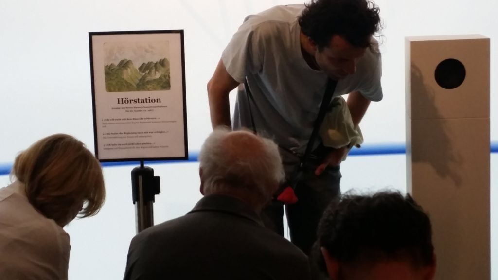 Neben einem hüfthohen Lautsprecher steht ein Mann, gebückt, und lauscht. Links von ihm steht eine Tafel, auf der Hörstation steht. Davor sitzen Leute, die man nur von hinten sieht.