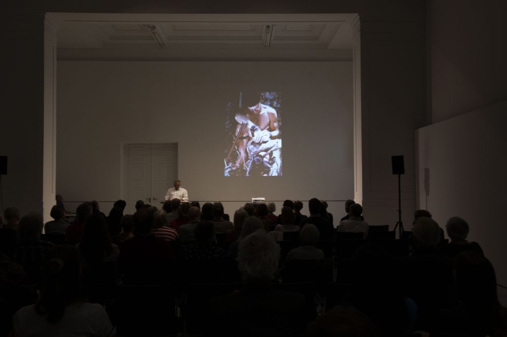 Ein dunkler Saal, in dem viele Leute sitzen und auf eine Wand blicken, auf der ein Bild von Bruno Manser gezeigt wird.
