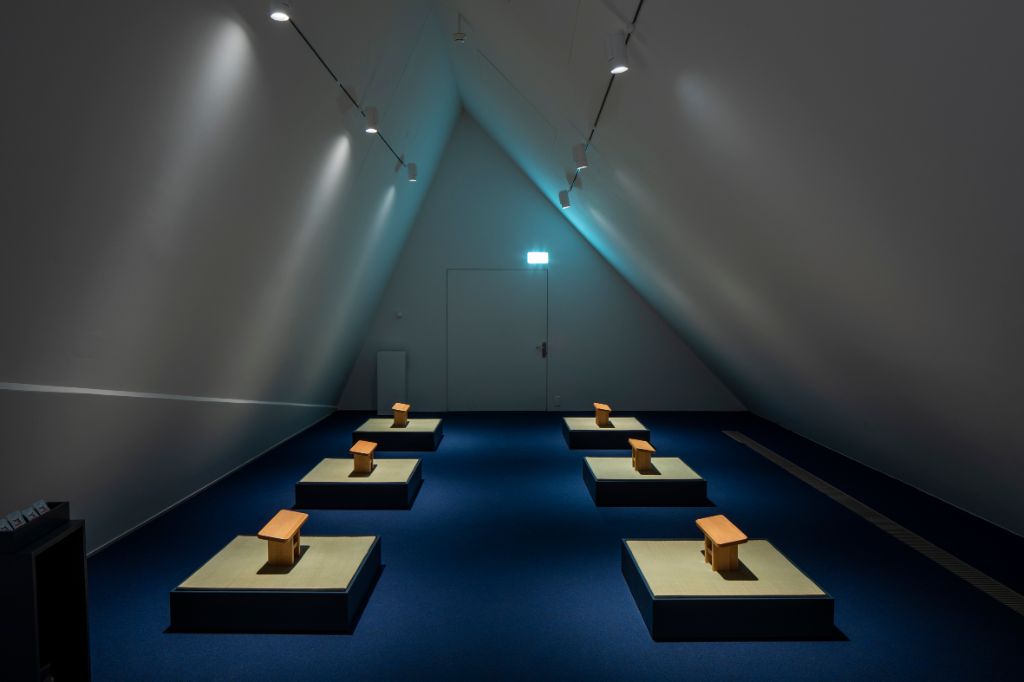 Inmitten der Ausstellung Erleuchtet hat es einen Meditationsraum, ausgestattet mit kleinen, hölzernen Hockern, die in einer Reihe, gegenübergestellt, aufgestellt sind.